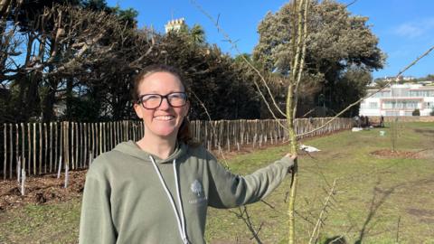 A white woman with brown hair, black glasses and a green hoodie holds a tree sapling and smiles at the camera. Behind her are a team of tree planters, planting hedgerows. 