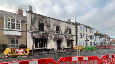 A burned out two-storey building, windows blown out and blackened, cream coloured front covered in smoke damage. Large wire fences and orange. barriers on the road on front.