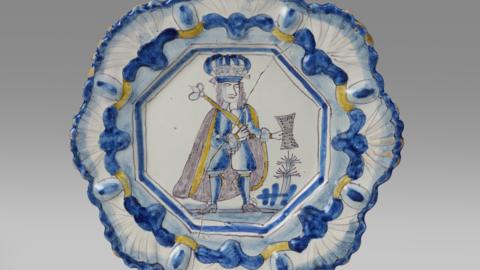 The yellow, blue and white plate depicts King James II holding a sceptre and parchment. The plate is against a white backdrop. The rim of the plate is moulding with a repeating shell motif.