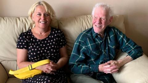 A woman with short blonde hair in a bob sits on a light-coloured sofa next to an older man. She wears a black dress with a small white flower pattern and holds a bright yellow handbag on her knee. The older man has white hair and wears a green checked shirt and khaki green trousers.