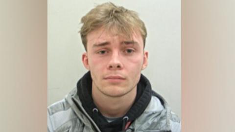 Matthew Bennett's police mugshot. He has blond hair, longer on top and shorter at the sides, and is wearing a black hoodie under a grey jacket against a pale grey background.