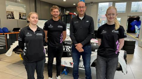 Alyce Mayo (left) and Kayley Philpott (right) standing in an engineering plant with the shell of a racing car behind them. They are stood with two men who are leading their course at the University of Wolverhampton. 