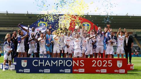 Leeds United players stand behind a banner saying Champions 2025 as they celebrate amid a shower of yellow and blue ticker tape.