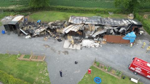 An aerial photograph shows a destroyed building with a charred and collapsed roof. Firefighters in beige and yellow uniforms can seen around the edged of the building. A red truck can be seen on the right.