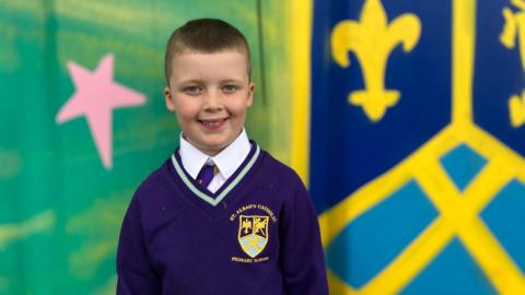Vinnie, who has short brown hair and wears a purple school uniform, smiles at the camera in front of a painted multi-colour mural.