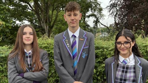Three young students in their school uniform smile at the camera as they stand in a line. The girl on the left has long brown hair. The boy has short brown hair while the girl on the right has long dark hair which is slightly curled and some of which has been tied back. They all wearing matching grey blazers with some yellow and purple details on them. The girls wear matching chequered grey and white pinafore dresses with white shirts underneath. The boy wears a white shirt under his blazer. They all wear matching grey, purple and blue ties.