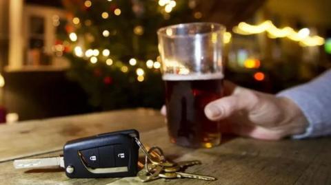A man holding a pint of beer, with car keys beside him and a Christmas tree in the background