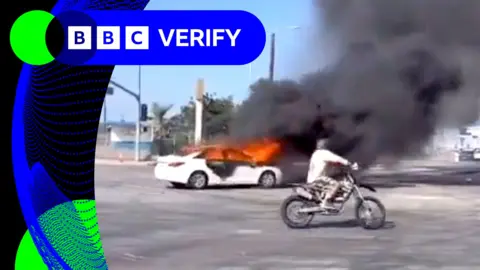 Car on fire with motorcycle driver in front.