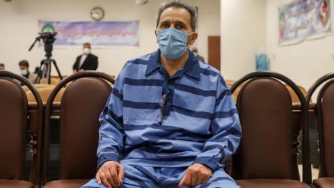 Jamshid Sharmahd has short greying hair and a blue medical mask across his face, as he sits in court wearing light blue prison prison fatigues with horizontal navy stripes. A video camera and banners are visible in the background during the hearing in Tehran, in February 2022