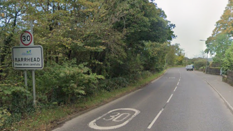 A stretch of road, surrounded by rural greenspace. A sign reads Barrhead and signals a speed limit change to 30mph