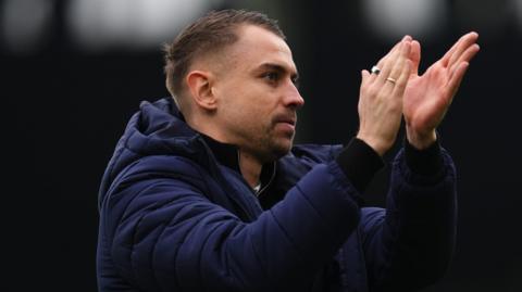 Southampton manager Tonda Eckert looks out with a focused look on his face and claps his hands in front of his face. He wear two silver rings on his right hand and a navy hooded puffer jacket.