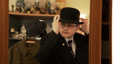 Charlie Elphinstone looks in a mirror to put on black bowler hat, he wears glasses, a white shirt, dark tie and suit and is surrounded by a military uniform and antique ornaments