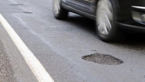 The bottom part of a car is seen, blurred as if it is moving along the road, which has a pothole in focus in the foreground and others seen further along the road.