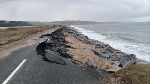The A379 Slapton Line between Torcross and Slapton. The road is damaged with one lane washed away. The sea is to the right and there is a grassy patch and a lake to the left.