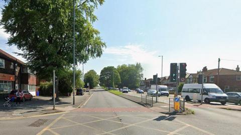 Daytime image of the A34 Kingsway in Manchester. There is no traffic on the left hand side of the road but several vehicles on the right. A woman holding a small child with a pram nearby stands on the pavement to the left of a yellow junction.