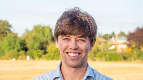 Cameron Holloway stares at the camera. He wears a blue and white checked shirt. In the background is a yellow field with green trees and hedgerows in the background. 