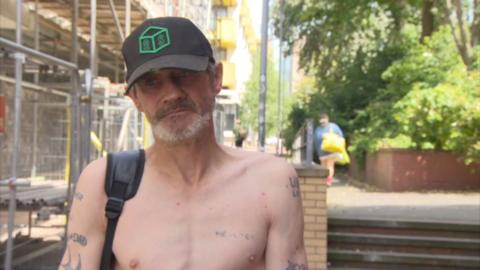 Ricky, with a grey beard and wearing a black baseball cap, stands near the spot where one of his friends died from a drugs overdose. There are tattoos on his bare chest, honouring friends he has lost