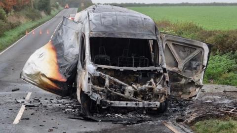 The van is parked at the side of the road. Much of it is black and clearly wrecked by the fire. A once white panel has been seemingly blown open by the fireworks on the left side of it, while the right seemed to have detached. 