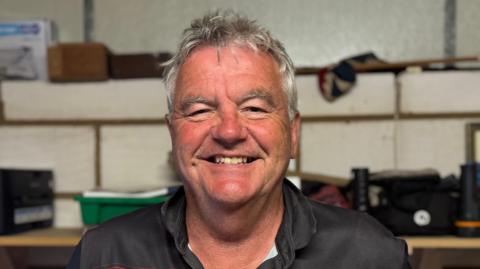 A head and shoulders image of Michael Hancock. He is wearing a black and red top and standing in front of a shelf that has various items on it. He is smiling and looking directly into the camera. 