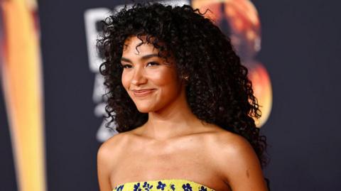 Olivia Dean is wearing a yellow strapless dress with navy blue flowers. Her hair is dark brown and curly, she has brown eyes. She is smiling slightly to the left of the camera. 