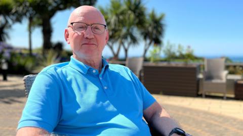 Mike is sitting outdoors on a sunny day, wearing a blue polo shirt and dark pants. The background includes palm trees, lush greenery, and outdoor furniture under a clear blue sky.
