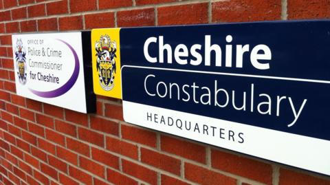 A Cheshire Constabulary headquarters sign that is blue and white with the force's badge next to it, set on a brick wall.