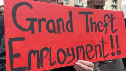 Banner from protestors outside Rockstar Games' offices reading 'Grand Theft Employment!' It's in black writing on a red background being held by what appears to be a woman's hand
