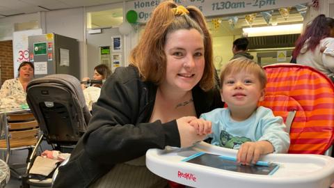 Kelly Kew wearing a green shirt and a black jacket- sitting next to her toddler in a high chair 