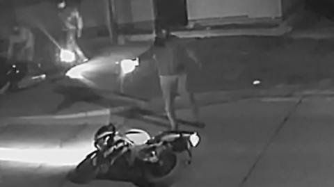 A man is standing over a motorbike which is led on its side on a residential road. He is pointing a gun and has shot it at the victim. Behind the motorbike is two other men.