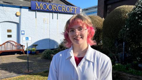 A girl with pink and blonde hair. She is looking at the camera smiling and is wearing a white overcoat with a red t-shirt underneath and silver rimmed glasses.