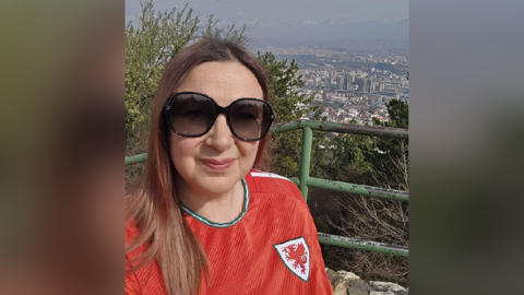 A woman in a red Wales shirt. She is wearing sunglasses and looking at the camera. It is a selfie and behind her is the view of a city. 