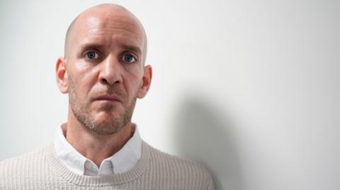 Dan Barr, 41, who has blue eyes, a bald head and light stubble, speaks into the camera wearing a beige jumper over a white shirt. 
