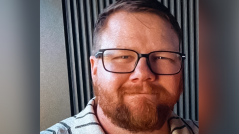 Mark Orpin in a family photo wearing a grey polo top with black stripes. He has shot cropped brown hair, an ginger beard beneath black rimmed glasses. He has closed mouth smile and has a kind face