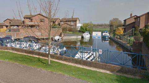 Riverside Mead, Peterborough, showing a piece of water, a bridge, boats on the water and houses either side of the river. There is a tree by the bridge. 