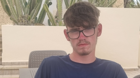 A boy with short brown hair, glasses and a blue shirt. There is a grey-coloured table and chair behind him and tall green cactus-type plants on top of a white wall.
