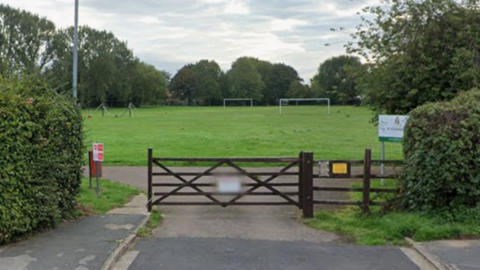 Google shot of an entrance to St Aiden's Park. There is a large wooden gate and football goals in the distance. There is also a red bin to the side of the gate.