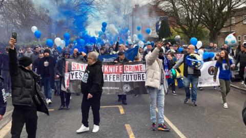 People on a street are letting off blue and white flares and balloons. Many are holding phones and taking selfies. One man is seen holding a wreath. Behind them a banner can be seen reading: "Say no to knife crime and yes to a safer future." 