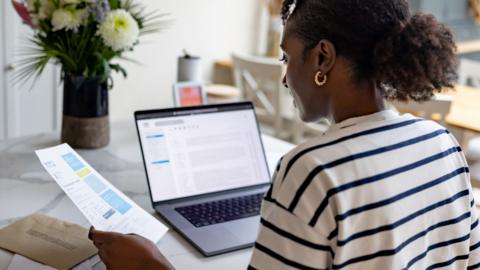 A woman looks at her bills in a letter and on a laptop whilst sat in her kitchen at home