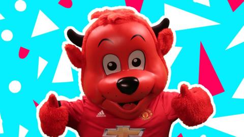 'Fred the Red' - official mascot of Manchester United FC. The mascot is red, has black horns and a large, smiling head. Wearing a MUTD football shirt and giving thumbs up.