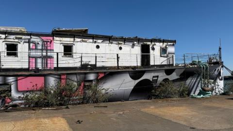 HMS President with grey and pink paintwork. The boat looks dilapidated and lots of weeds are growing up its sides.
