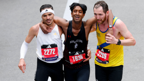 Three men wearing black shorts and different coloured running vests jog towards the finish line of the Boston Marathon with their arms around each other. Robson De Oliveira, who has short, dark hair and a moustasch and is wearing a white vest and a white headband. Ajay Haridasse has short, dark hair and a black vest. Aaron Beggs has short brown hair and a beard and is wearing a yellow vest with a blue stripe.