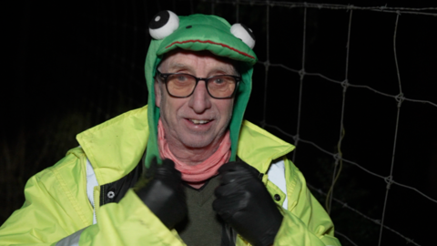 A man wearing a felt hat in the shape of a frog and dark-rimmed glasses, he has a hi-vis jacket and black leather gloves. 