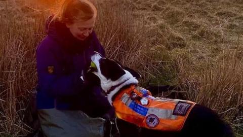 A woman wearing waterproofs playing with a collie dog wearing an orange fluorescent jacket in a field, with the sun setting behind them