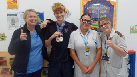 Three female hospital staff standing with a 15-year-old boy. The women are all in scrubs, two of them in grey and one in light blue. Two of them have their thumbs up while the third is standing with her arms at her side. All of them are looking at the camera and smiling. The boy has short, curly blonde hair and is wearing dark clothing with two medals round his neck. He is holding them, looking at the camera and smiling.