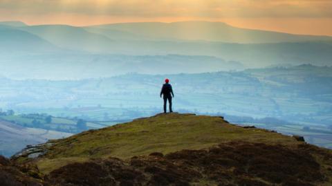 A person is standing on top of a mountain during sunset looking down on roaming fields.