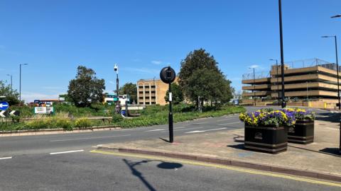 A roundabout viewed from one of the approaching roads. In the distance are buildings, including a multi-storey car park.