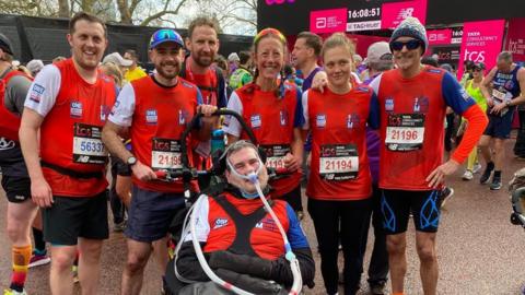 Group stood at the finish of a sporting event, with six people in matching red tops stood with Sam Perkins, who is in a wheelchair.