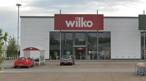 A large retail unit at an out-of-town retail park. There is a large rectangular sign saying wilko above the glass entrance. Two cars are parked in the car park in front of the building.