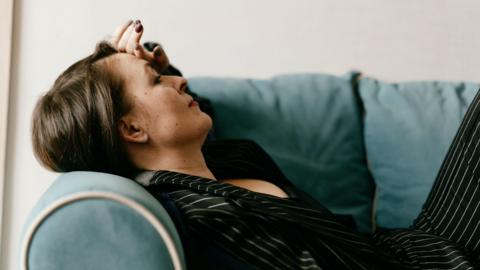 A woman lays on a sofa arm looking fatigued.