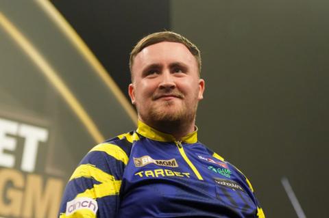 Luke Littler has short brown hair and wears a yellow and blue darts top. He is smiling.